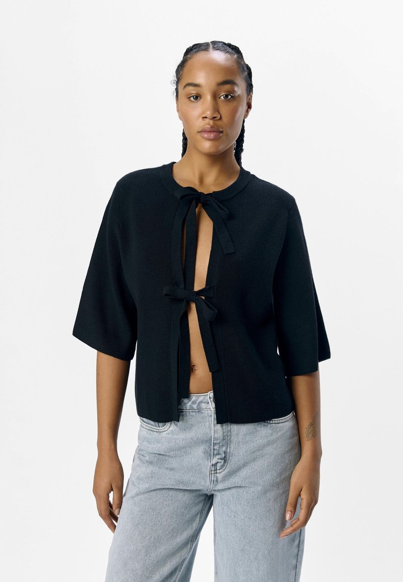 Schwarzer, gestrickter Cardigan mit kurzen Ärmeln, der einen Knotenverschluss am Hals und ein leicht offenes Design vorne aufweist. Getragen mit hellblauen Jeans.