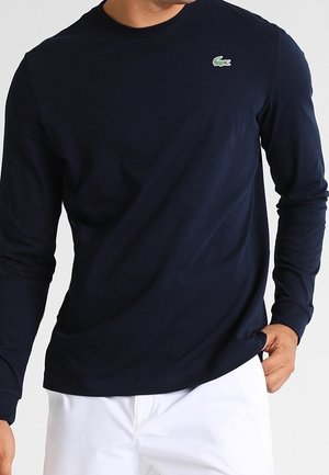 Homme portant une chemise bleu marine à manches longues avec un petit logo crocodile vert sur la poitrine et un pantalon blanc, la main tenant l'ourlet de la chemise.