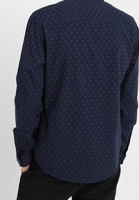 Marineblauwe blouse met lange mouwen en kleine, herhalende tonale patronen. Het materiaal lijkt glad met een standaard kraag en knopenmanchetten.