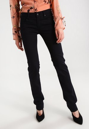 Femme portant un pantalon noir ajusté, des escarpins noirs à bouts pointus, et un chemisier à manches longues à motif floral pêche avec des détails de boutons.