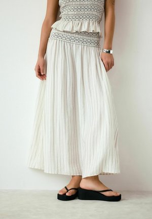 Kvinde iført lang hvid og gråstribet nederdel, tekstureret ærmeløs top, sorte platformsandaler og et sølvarmbånd på hendes højre håndled.