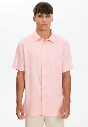 Joven con cabello corto y oscuro, vestido con una camisa de manga corta color rosa claro abotonada y pantalones beige sobre un fondo blanco.