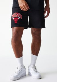 Calções de basquetebol em malha preta com o logótipo "Chicago Bulls" vermelho na perna esquerda. Combinados com ténis brancos e meias de cano curto.
