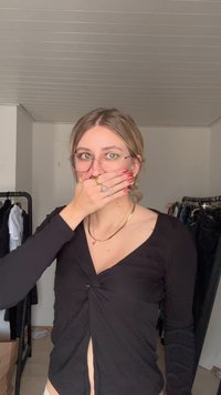 Top preto de manga comprida com design torcido na frente e corte ligeiramente aberto. Acessórios incluem colares dourados em camadas e anéis.