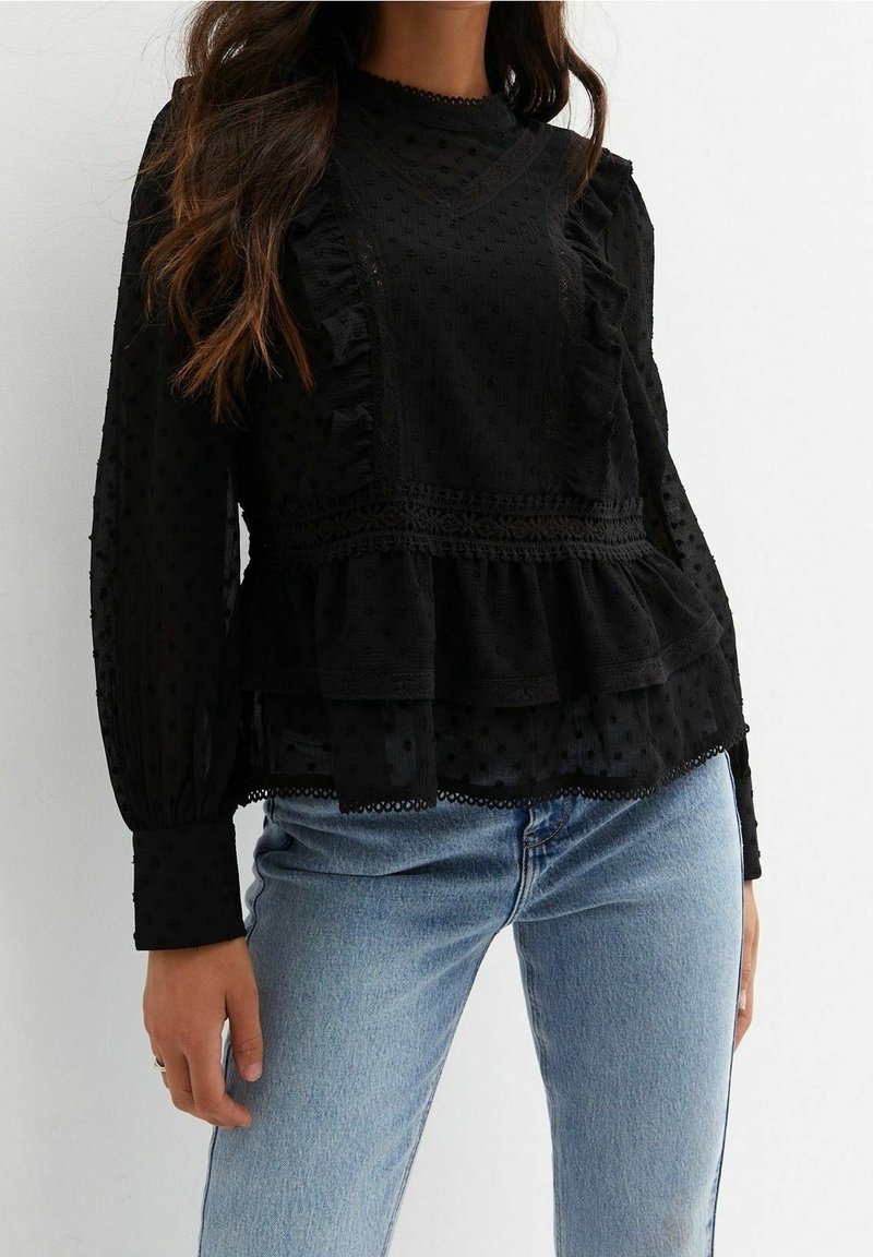 Blusa nera con texture, dettagli a volant e accenti in pizzo. Maniche lunghe con polsini arricciati, orlo peplum a strati, indossata sopra jeans blu.