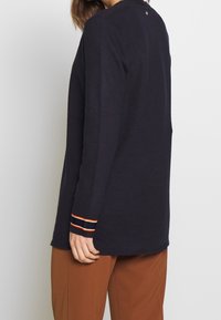 Marineblaues Langarmoberteil mit Rundhalsausschnitt. Verfügt über gestreifte Bündchen in Rot und Weiß. Leichtes Strickmaterial mit lockerer Passform.