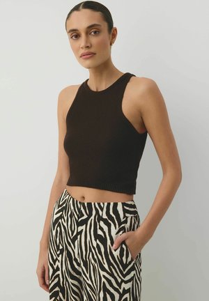 Kobieta w czarnym, bezrękawnym topie do pasa i wysokich spodniach w zebra wzór, z jedną ręką w kieszeni na jednolitym tle.