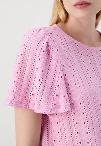Różowa bluzka z haftowanym wzorem w oczka, falbanastymi rękawami i okrągłym dekoltem. Materiał ma teksturową, przewiewną jakość.