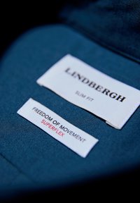 Blauwe stoffen label met twee witte etiketten: "LINDBERGH" en "Slim Fit" boven "VRIJHEID VAN BEWEGING" en "SUPERFLEX." Tekst in zwart en rood.