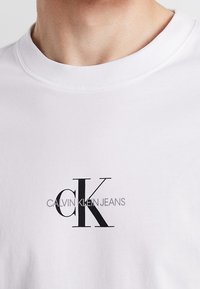 Valkoinen puuvillapaita, jossa on pyöreä kaula-aukko ja lyhyet hihat, musta "CK" -logo ja teksti "CALVIN KLEIN JEANS" alla.