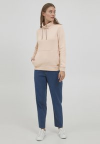 Felpa rosa chiaro con colletto alto, cordino regolabile, tasca frontale e spacchi laterali, abbinata a pantaloni in denim blu e sneakers bianche.