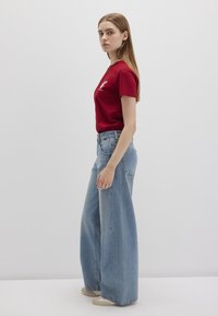 Red cotton t-shirt with short sleeves and graphic detail, paired with high-waisted blue wide-leg jeans. Model stands sideways against a plain background.