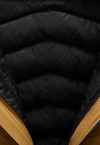 Veste à manches longues avec un intérieur matelassé noir et un extérieur en tissu couleur miel, mettant en valeur une fermeture éclair élégante et un motif en chevrons.