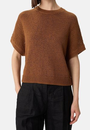 Femme portant un pull en maille marron à manches courtes avec des manches texturées et un pantalon noir sur un fond uni.
