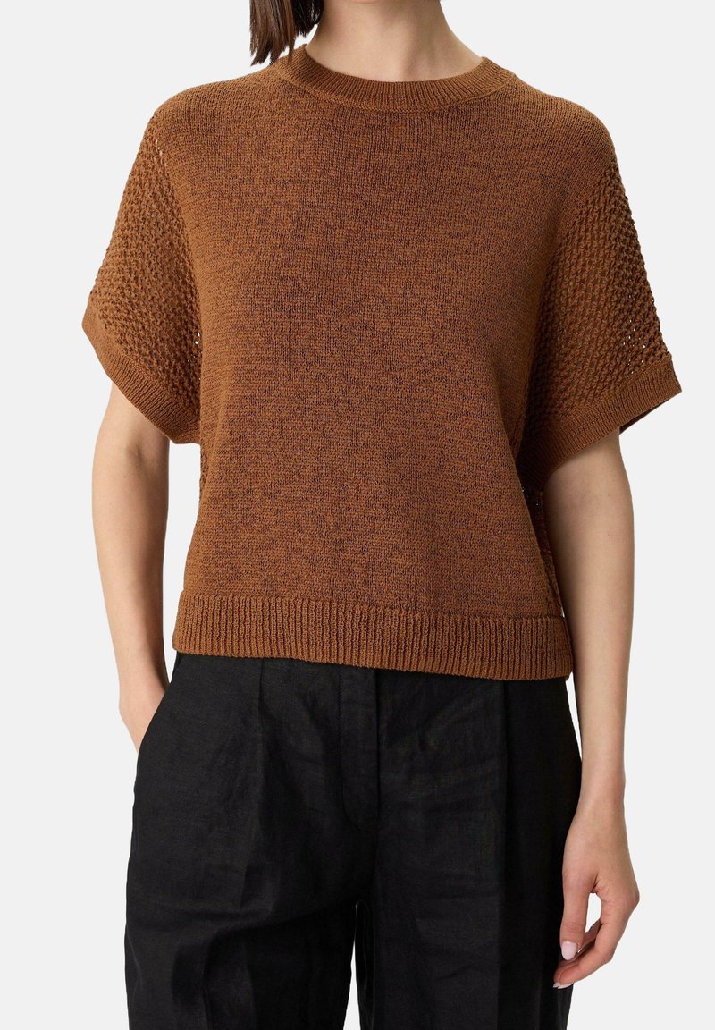 Femme portant un pull en maille marron à manches courtes avec des manches texturées et un pantalon noir sur un fond uni.