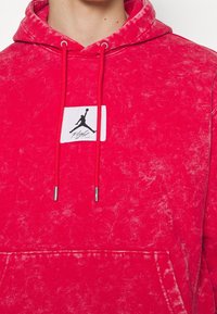 Sudadera roja con un acabado texturizado, que presenta un parche blanco con un logo de silueta negra. Los cordones son rojos con puntas metálicas.