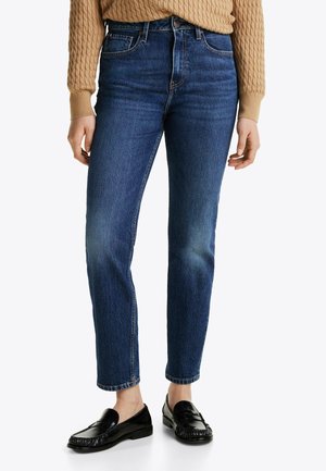 Osoba ubrana w niebieskie dżinsy o prostych nogawkach, czarne mokasyny i beżowy sweter z grubym splotem, stojąca na jednolitym tle.