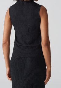 Haut sans manche noir en tissu texturé avec un col montant et une coupe ajustée, faisant partie d'un ensemble assorti. Présente une texture côtelée subtile.