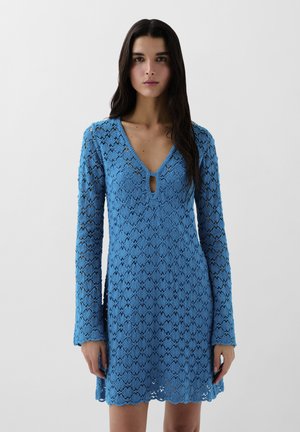 Rochie albastră din dantelă cu mâneci lungi, de tip V, și detaliu cu deschidere. Prezintă un tiv ondulat și un model texturat pe întreaga suprafață.