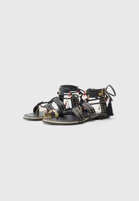 Sandali in pelle nera con borchie dorate, multiple cinghie, charm decorativi e una frangia nera. Suola piatta e design open toe.