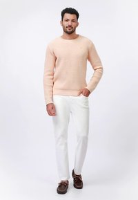Maglione di colore arancione chiaro con trama, scollo rotondo, abbinato a pantaloni bianchi e scarpe marrone scuro. Piccolo logo decorativo sul maglione.