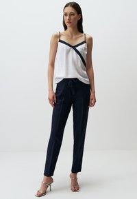 Witte camisole met marineblauwe details, gecombineerd met marineblauwe broek met een trekkoord in de taille. Zilveren sandaaltjes met bandjes geven de outfit de finishing touch.