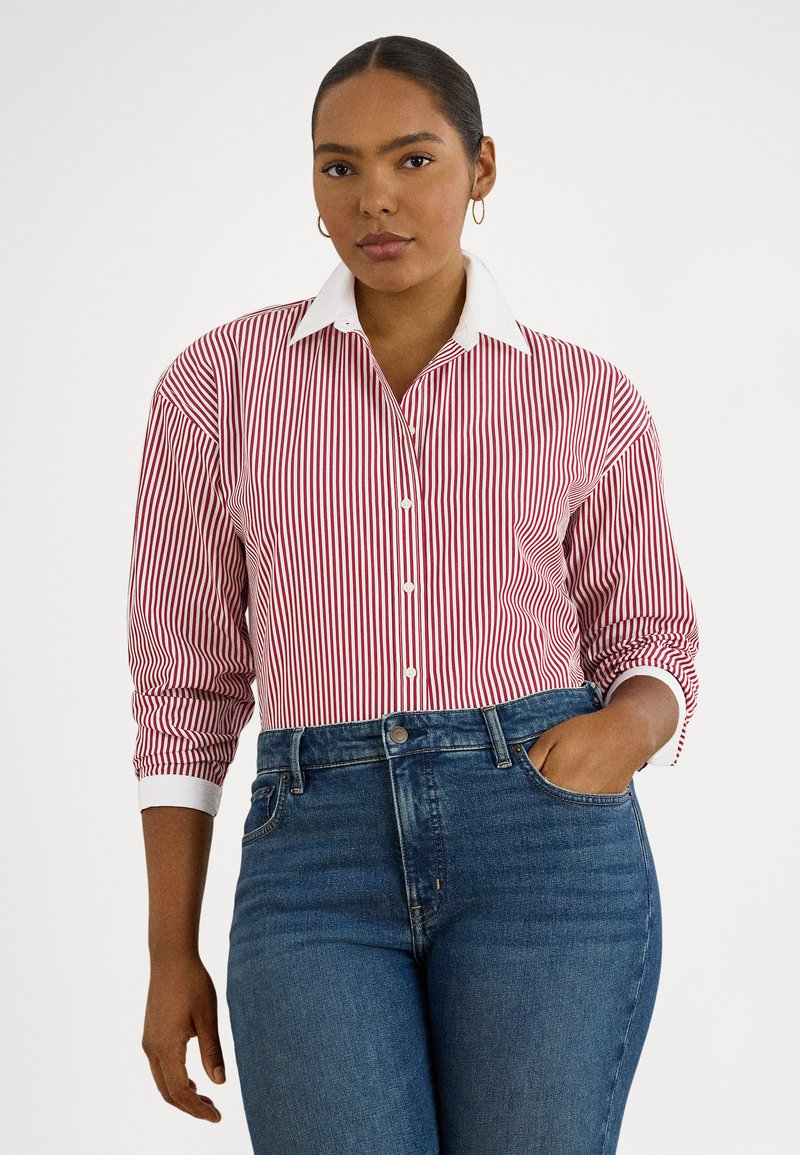 Γυναίκα με σκούρα μαλλιά μαζεμένα πίσω, φορά χρυσούς κρίκους στα αυτιά, κόκκινο και άσπρο πουκάμισο με κάθετες ρίγες, λευκό γιακά και μανσέτες, και μπλε τζιν.