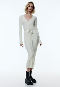 Langes cremefarbenes, geripptes Strickkleid mit tiefem V-Ausschnitt, Knopfleiste und Bindegürtel. Kombiniert mit klobigen schwarzen Stiefeletten.