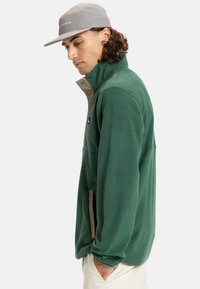 Giacca in pile verde con colletto alto, dettagli in zip beige e tasche laterali. Il materiale è morbido e testurizzato, offrendo calore.