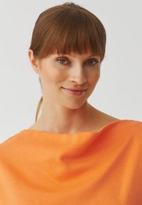 Top arancione, leggero, con una texture morbida, caratterizzato da un ampio scollo e un design fluido che si adagia dolcemente sulle spalle.