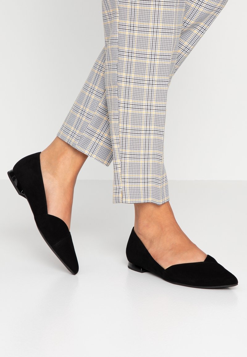 Zapatos planos de ante negros con punta afilada y lados cortados, complementados con un tacón bajo. Llevados con pantalones de cuadros en beige, azul y amarillo.