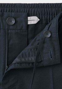 Gros plan d’un pantalon noir avec fermeture éclair partiellement ouverte et bouton, taille élastique et patte latérale réglable.
