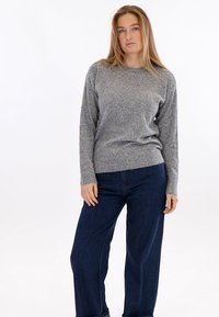 Jonge vrouw met lang blond haar, gekleed in een grijze trui en donkerblauwe spijkerbroek, staand tegen een effen witte achtergrond.