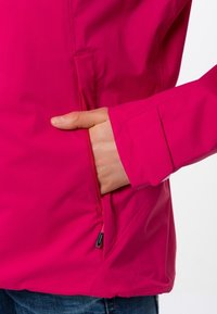 Una mano inserita nella tasca con zip di una giacca rosa brillante indossata sopra dei jeans blu su uno sfondo bianco.