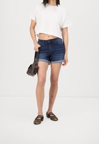 Mujer vestida con una camiseta blanca corta, pantalones cortos de mezclilla oscuros, zapatos planos de print de leopardo y sosteniendo un bolso marrón oscuro por la correa.