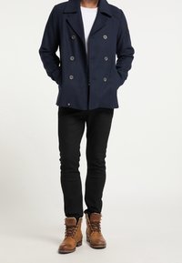 Tmavě modrý dvouřadý kabát z vlněné směsi s velkými knoflíky, doplněný černými přiléhavými kalhotami a hněd�ými koženými botami na šněrování.