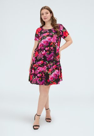 Mujer que lleva un vestido negro con estampado floral en tonos rosa brillante y rojo, mangas cortas y sandalias negras de tacón, de pie con una mano en la cadera.