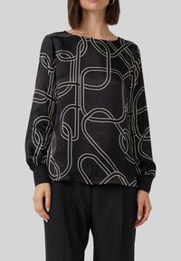 Schwarze Bluse mit langen Ärmeln aus glattem Stoff, die ein geometrisches weißes Muster aus Schlaufen aufweist. Die Manschetten sind tailliert und der Ausschnitt ist weit.