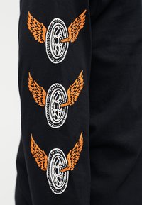 Maglietta nera a maniche lunghe con tre grafiche ripetute di una ruota con ali arancioni, dettagliata con un contorno puntinato attorno alla ruota.