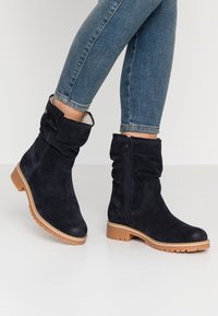 Botas de tobillo de ante azul marino oscuro con un eje caído, cremallera lateral y suela de goma beige. Cuenta con un puño plegable en la parte superior.