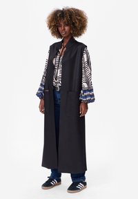 Long manteau noir sans manches avec deux poches avant, porté sur une chemise à motifs avec des accents bleus et un jean en denim, associé à des baskets.