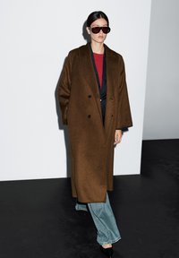 Brązowy wełniany płaszcz o podwójnym zapięciu, w zestawieniu z czerwonym topem i niebieskimi szerokimi dżinsami, dopełniony błyszczącymi czarnymi butami i dużymi okularami przeciwsłonecznymi.