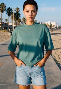 Jeune personne aux cheveux courts et bouclés portant un t-shirt bleu sarcelle et un short en jean, debout sur un chemin ensoleillé sur la plage avec des palmiers et des bâtiments en arrière-plan.