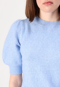 Kvinna i en ljusblå kortärmad tröja med ribbad rundhals och puffärmar mot en enfärgad vit bakgrund.