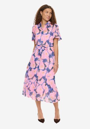 Kobieta w różowej sukience midi w kwiaty z krótkimi rękawami, z przodu zapinanej na guziki, z paskiem w talii i czarnymi szpiczastymi szpilkami, stoi na białym tle.