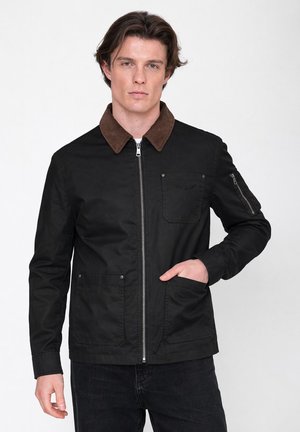 Junger Mann trägt eine schwarze Jacke mit braunem Kragen, Frontreißverschluss, mehreren Taschen und schwarze Hose, steht vor einem weißen Hintergrund.