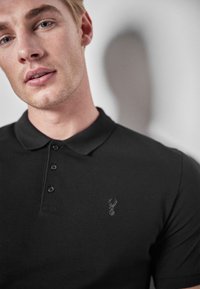 Sort polo-skjorte lavet af tekstureret stof, med en tre-knaps placket og et subtilt broderet logo på venstre bryst.