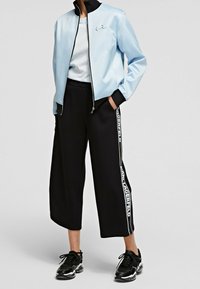 Helle blaue Bomberjacke mit schwarzen Akzenten, weißes T-Shirt, schwarze weit geschnittene Hose mit seitlichem Logo und schwarze Sneaker.