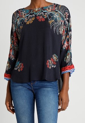 Femme portant un chemisier noir à manches longues avec des motifs floraux colorés sur les manches et la poitrine, assorti d'un jean bleu.