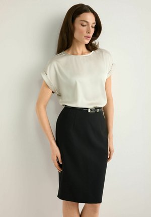 Kobieta w kremowej bluzce z krótkimi rękawami, wpuszczonej w czarną ołówkową spódnicę z wysokim stanem, przewiązaną wąskim czarnym paskiem, stojąca na tle jednolitej ściany.